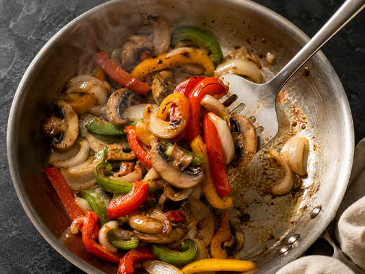 Sautéed peppers, onions and mushrooms in a stainless steel pan being stirred with a steel spatula — preventing food from sticking